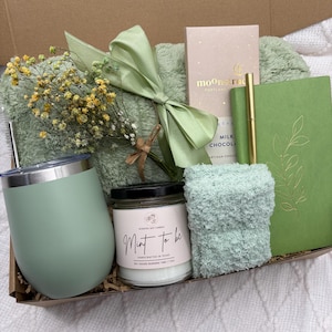 May include: A gift box filled with mint-colored items, including a tumbler, candle, socks, a journal, a pen, a chocolate bar, and a small bouquet of yellow flowers. The candle reads "Mint to be". The items are arranged on a green blanket.