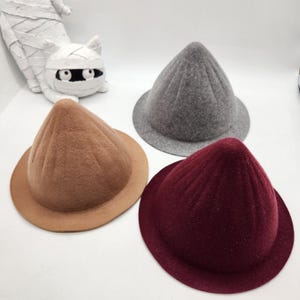 May include: Three felt hats in brown, gray, and burgundy. Each hat has a conical crown and a wide brim. The hats are displayed on a white surface. A white mummy cat figurine is in the background.