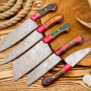 Op de afbeelding: Vijf Damascus stalen messen met kleurrijke houten handvatten. De handvatten hebben rode, blauwe, groene en gele accenten. De messen hebben een uniek patroon. De messen zijn gerangschikt op een houten oppervlak.