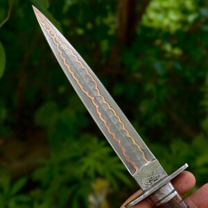May include: A handcrafted dagger with a long, pointed blade. The blade features a detailed pattern with copper-colored accents. The handle is made of wood and metal. The dagger is held against a blurred green background.