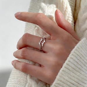 May include: A silver ring with a branch design. The ring has a delicate, openwork pattern resembling twigs or antlers. The ring is worn on a finger, highlighting its intricate details and minimalist style.