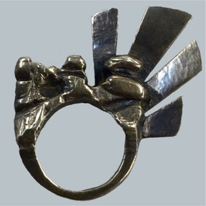 Könnte beinhalten: Ein silberner Ring mit einem strukturierten, abstrakten Design. Der Ring hat eine breite Band und eine große, unregelmäßige Form. Das Design verfügt über eine Reihe von scharfen, eckigen Punkten, die einen dramatischen Effekt erzeugen.