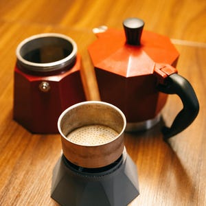 Peut inclure: Une cafetière italienne rouge et noire démontée. Les composants comprennent la base, le filtre à café et la chambre supérieure. La cafetière est en métal et possède une poignée noire. Les composants sont disposés sur une surface en bois.