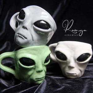 May include: Three ceramic mugs shaped like alien heads, in white and green. The mugs have large, black oval eyes and a detailed facial structure. The mugs are stacked on a dark, textured surface. The text "Ramya Ceramics" is in the upper right corner.