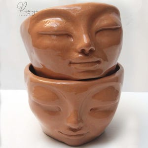 Puede incluir: Dos macetas de cerámica apiladas de color terracota. Cada maceta presenta un rostro sereno con los ojos cerrados, una nariz pequeña y una suave sonrisa. Las macetas son lisas y brillantes, con el nombre de la marca "Ramya Ceramics" en cursiva.