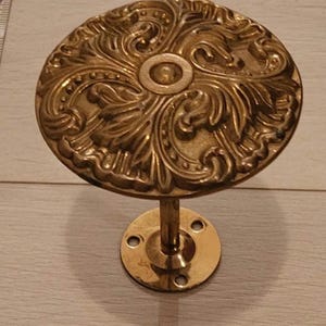 May include: Ornate brass curtain rod holder with a circular, floral-patterned design. The holder features a round base with three screw holes and a central stem supporting the decorative top. A vintage-style home decor item.