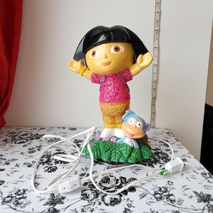 May include: A Dora the Explorer lamp featuring a smiling Dora figure with outstretched arms, wearing a pink shirt and a yellow bracelet. A blue monkey figure is also present. The lamp has a black base and a white power cord.