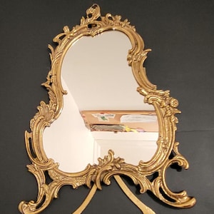 Peut inclure: Miroir de coiffeuse doré orné, de forme ovale. Le cadre est orné de volutes et de motifs décoratifs. Il est soutenu par deux pieds incurvés. Le design est vintage et décoratif.