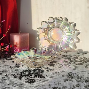 Peut inclure: Deux plats en verre irisé avec un bord festonné. L'un est rond, l'autre est un bol peu profond en forme de fleur. Une bougie rose et un rideau rouge sont en arrière-plan. Les plats reflètent la lumière, créant un effet arc-en-ciel.
