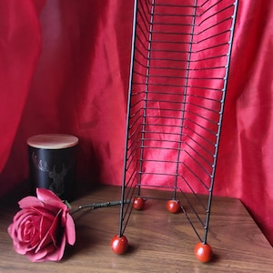 May include: A black metal magazine rack with a red rose and a black candle holder with a deer silhouette. The rack has a series of horizontal bars and rests on four red spheres.