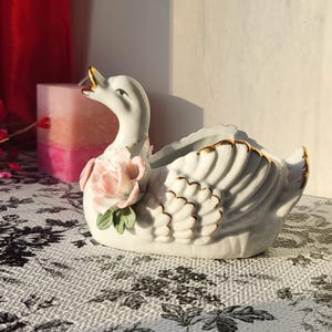 May include: A white ceramic swan planter with gold trim and a pink rose accent. The swan has a long neck and open beak, with detailed feathering on its wings. The planter is on a floral patterned surface.