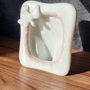 May include: A white ceramic photo frame with a pink scalloped edge and a dove detail. The frame has a rectangular shape with curved corners and is on a wooden surface.