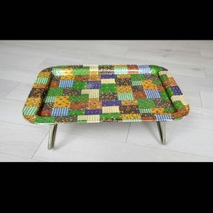 Può includere: Un vassoio rettangolare con un motivo patchwork nei toni del verde, giallo, blu e marrone. Il vassoio ha angoli arrotondati ed è sostenuto da gambe in metallo argentato. Il vassoio è lungo circa 51 cm e largo 36 cm.