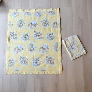 Peut inclure: Couverture de bébé jaune et bavoir assorti. La couverture présente un motif répétitif d'illustrations de dessins animés en bleu, vert et marron. La couverture a un bord à volants.