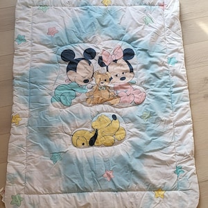 Könnte beinhalten: Eine rechteckige Babydecke mit weißem Rand, die Micky Maus, Minnie Maus und Pluto zeigt. Die Decke hat einen hellblauen und weißen Wolkenhintergrund. Micky und Minnie sind mit einem Teddybär dargestellt, und Pluto liegt.
