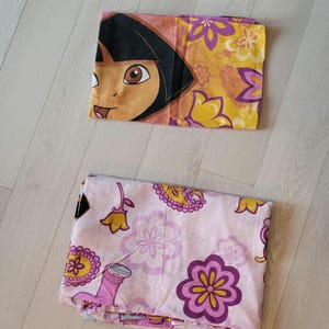 Peut inclure: Deux taies d'oreiller pliées. L'une présente un personnage de dessin animé aux cheveux noirs et aux yeux bruns sur un fond floral jaune et orange. L'autre est rose avec des motifs floraux violets et jaunes. Les taies d'oreiller sont sur un sol en bois clair.