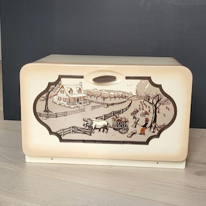Puede incluir: Panera crema con un panel decorativo que representa una escena rural en tonos marrón y beige. La escena incluye una casa, árboles, una valla y figuras con un carro y bueyes. La panera tiene un asa en la parte superior.