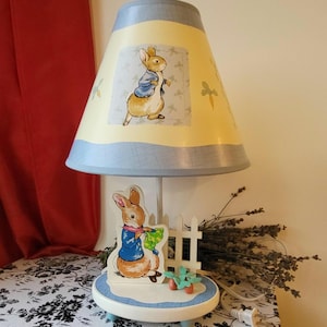 May include: A charming table lamp with a Peter Rabbit design. The lampshade is yellow with a blue border and a rabbit illustration. The base features a rabbit figure and a small white picket fence. Ideal for a child's bedroom, it adds a touch of whimsy.