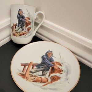 Peut inclure: Un ensemble tasse et assiette en céramique blanche avec une scène nautique. L'illustration représente une personne en manteau bleu sur un radeau, naviguant dans des eaux agitées. L'assiette et la tasse ont une bordure dorée.