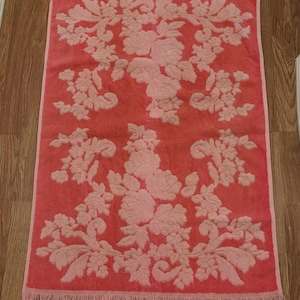 May include: A rectangular coral-coloured bath towel featuring a floral pattern in a lighter shade of pink. The towel has a fringed edge and is on a wooden floor. The floral design is symmetrical, with large blooms and detailed leaves.