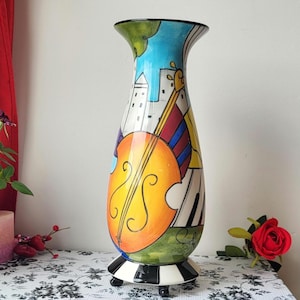 May include: A tall, hand-painted ceramic vase with a musical theme. The vase features a cello, piano keys, and a cityscape design in vibrant colours. The base has a black and white checkerboard pattern, and the vase is approximately 30 cm tall.