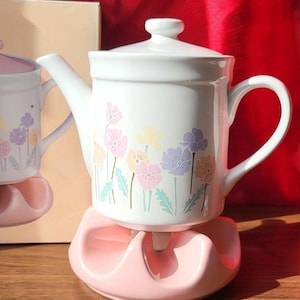 May include: A white ceramic teapot with a lid, decorated with pastel floral patterns. It rests on a pink ceramic warmer. The teapot features a curved handle and spout. The design includes pink, yellow, and purple flowers.