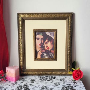 May include: Framed art print featuring two women with red lipstick and dark hair. The artwork is in a gold ornate frame with a cream-colored mat. A pink and peach candle and a red rose are on a table with a black and white floral pattern.