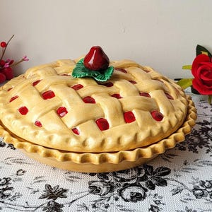 Puede incluir: Una fuente de cerámica para pastel con un diseño de corteza de rejilla. El pastel es de color canela con detalles rojos, coronado con una fresa roja y hojas verdes. El plato tiene un borde festoneado.