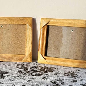May include: Two rectangular wooden picture frames with a light brown finish. The frames are empty, with a cardboard backing. A red rose with green leaves rests on the patterned surface next to the frames. The frames are on a table.