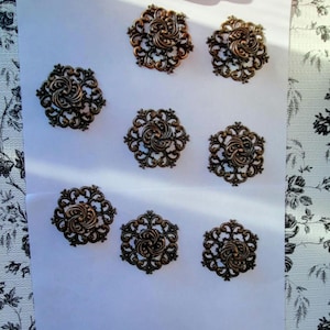 Puede incluir: Nueve botones decorativos de color bronce con un diseño de filigrana floral ornamentado. Cada botón presenta una espiral central rodeada de delicados patrones calados. Los botones están dispuestos sobre una superficie blanca.