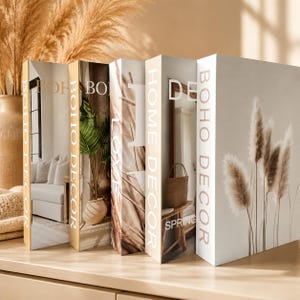 Peut inclure: Cinq livres décoratifs avec les mots "BOHO DECOR" imprimés sur les dos. Les couvertures des livres présentent des images d'intérieurs et d'herbe de la pampa. Les livres sont disposés sur une surface en bois de couleur claire.