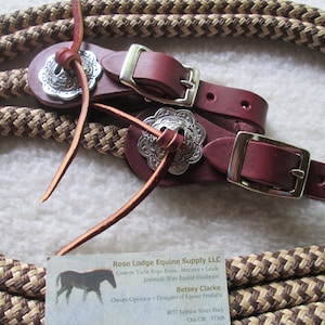 May include: Two brown leather reins with silver conchos and buckles. The reins are made of braided rope and are designed for horses. The reins are on a white background with a business card that reads "Rose Lodge Equine Supply LLC" and "Betsey Clarke, Owner/Operator & Designer of Equine Products".