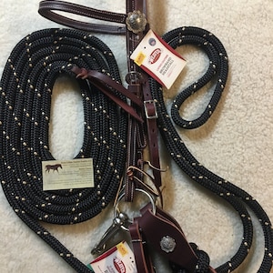 May include: A horse bridle and rope set. The bridle is dark brown leather with silver-colored accents. A black rope with tan flecks is coiled and looped. Weaver brand tags are attached.