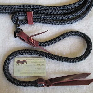 May include: Black braided rope halter with brown leather accents and a black snap hook. The halter is coiled on a white surface. The text on the card reads "Rose Lodge Equine Supply LLC, Custom Tack & Rope Halters, Made in USA,  Betsy Clarke, Unique Designs of Western Products, Natural Horsemanship Training and Trail Equipment, 807-337-7048".