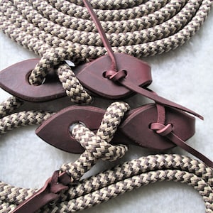 May include: A brown and tan braided rope with leather ends. The rope is tied in a knot with the leather ends forming a loop. The rope is coiled up in the background.
