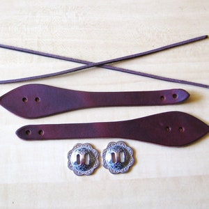May include: Two dark brown leather straps with pointed ends and two pre-punched holes, two thin leather cords, and two silver-toned metal conchos with a floral-inspired scalloped edge and a central rectangular slot for attachment.