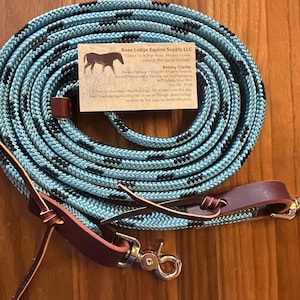 May include: A coiled light blue and black braided rope lead with dark brown leather accents and a silver metal swivel snap hook. A business card for Rose Lodge Equine Supply LLC featuring a horse silhouette graphic is placed on the rope.