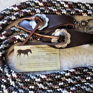 May include: Equestrian rope reins with dark brown leather accents and silver-toned decorative conchos. The rope is a mix of white, brown, and black. The reins have a clip for easy attachment. A business card is visible.