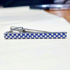 Puede incluir: Un pasador de corbata plateado con un patrón de cuadros azules y blancos. El pasador es rectangular con un diseño articulado. El patrón de cuadros es una cuadrícula repetitiva de cuadrados azules y blancos.