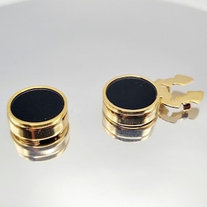 Peut inclure: Deux boutons de manchette dorés avec un centre circulaire noir. Un bouton de manchette a un fermoir doré. Les boutons de manchette sont sur une surface réfléchissante. Les boutons de manchette sont probablement vintage ou antiques.