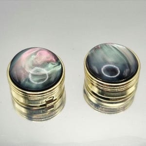 以下が含まれることがあります： 2つの小さく丸い金色の箱で、虹色と真珠色の蓋が付いています。蓋にはピンク、緑、グレーの渦模様があります。箱はヒンジ付きのデザインで、ジュエリーや薬など、小さなアイテムの保管に使用される可能性があります。