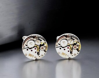 Puede incluir: Un par de gemelos circulares que muestran movimientos de reloj mecánicos expuestos con engranajes plateados, pequeñas joyas rojas y detalles dorados. Los componentes metálicos tienen grabadas las letras LSE en una fuente clara.