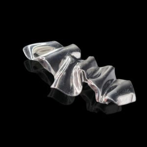 May include: A silver-colored, abstract metal sculpture with a rippled, flowing design. The reflective surface creates highlights and shadows against the black background. The piece appears to be a decorative object.