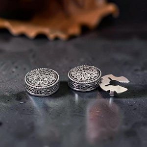 May include: Two circular silver-toned metal buttons featuring intricate embossed patterns on the top surface and a braided border. One button is shown from the side with a metal attachment piece extending from its base, while the other is flat.