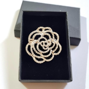 Peut inclure: Broche dorée en forme de fleur ornée de cristaux clairs et étincelants. La broche est présentée dans un coffret noir avec un intérieur en velours noir et un couvercle gris. Le design floral est détaillé.