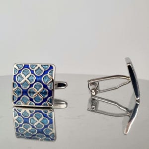 Op de afbeelding: Een paar vierkante manchetknopen met een blauw en lichtblauw bloemmotief op een zilveren basis. De manchetknopen worden gepresenteerd op een reflecterend oppervlak, dat hun ontwerp en vakmanschap laat zien.