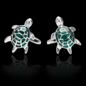 May include: A pair of silver cufflinks in the shape of sea turtles. Each turtle has a green, patterned shell and outstretched flippers. The cufflinks are set against a black background.