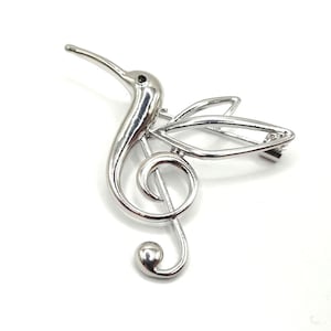 Pode incluir: Broche prateado em forma de beija-flor combinado com uma clave de sol. O beija-flor tem um bico longo e um pequeno olho preto. As asas estão abertas, e a clave de sol forma o corpo e a cauda do pássaro. O broche tem um fecho de alfinete.