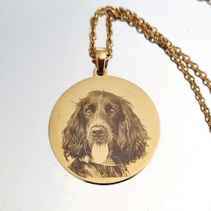 Puede incluir: Collar con colgante dorado con un retrato grabado de un perro. El perro tiene pelo largo y ondulado y la lengua fuera. El colgante circular cuelga de una cadena dorada. El colgante es de metal.