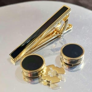 以下が含まれることがあります： 黒いエナメルインセットが付いた、金色のネクタイクリップと2つのマッチングカフスボタン。ネクタイクリップは長方形で、カフスボタンは丸いです。金色の装飾要素も存在します。